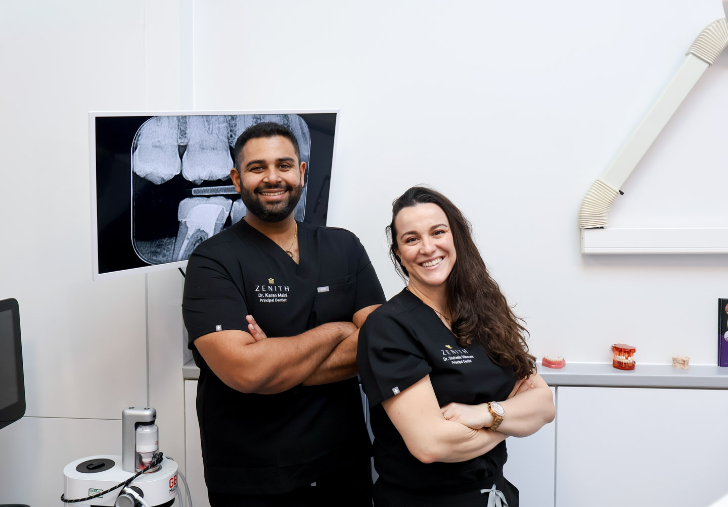 Two Zenith dentists in black scrubs smiling with arms crossed in a dental surgery, a dental X-ray displayed on screen behind them