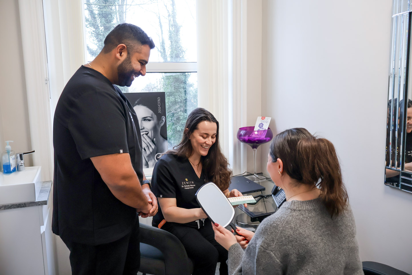 Two dentists in black scrubs smiling with a patient holding a hand mirror during a teeth whitening consultation at Zenith Dental