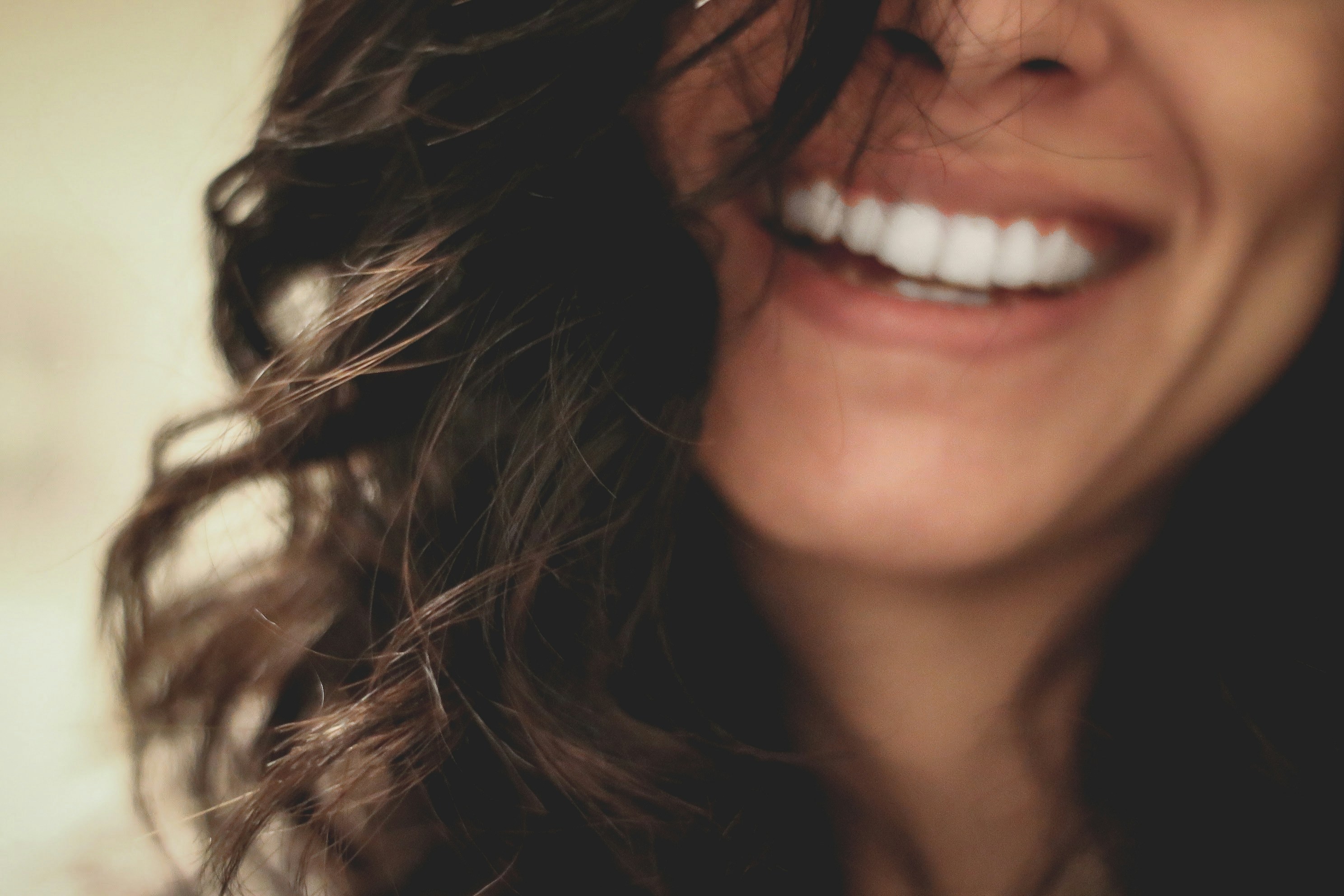 Close-up of a woman with dark curly hair laughing, showing bright white teeth against a soft blurred background