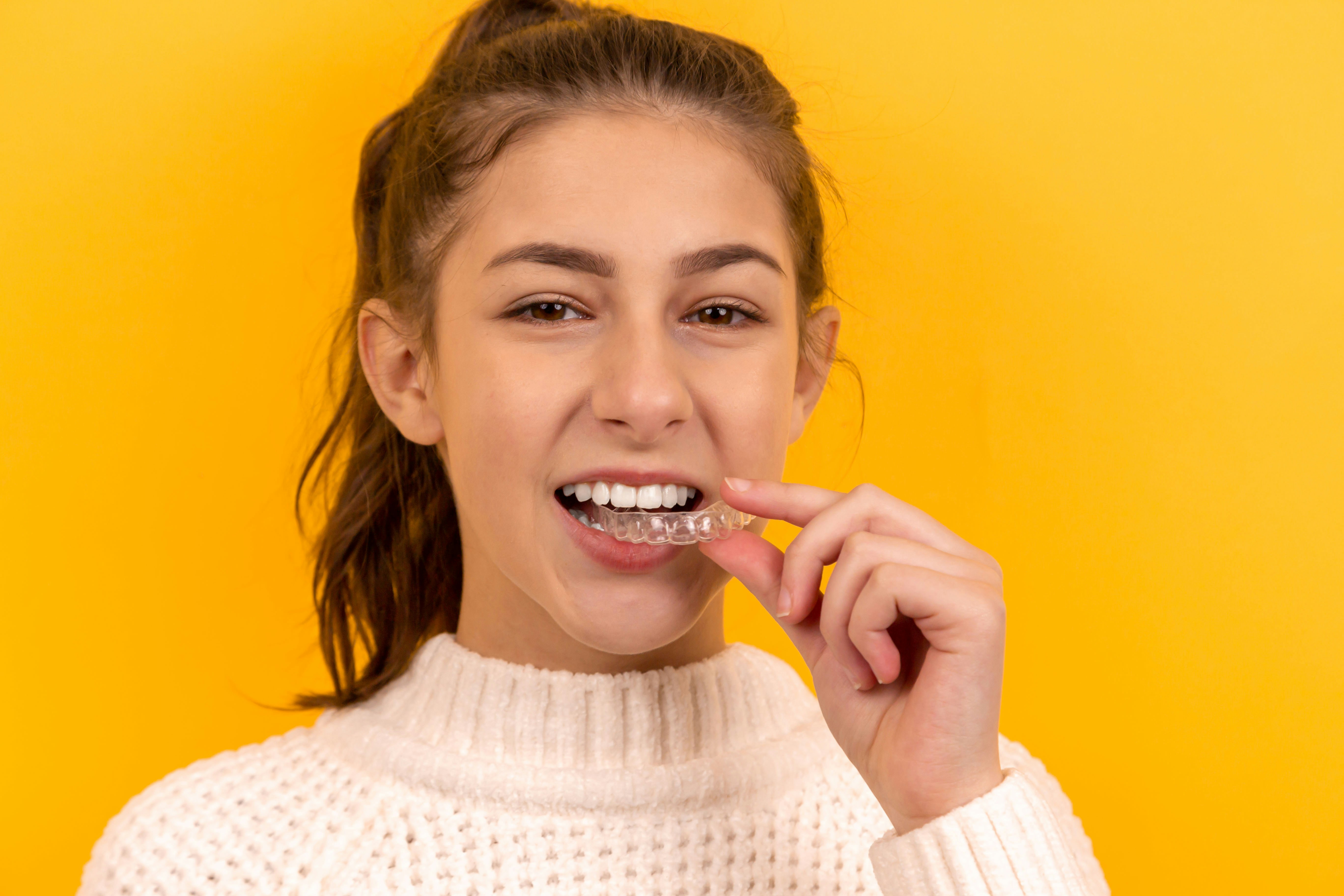 Smiling young woman inserting a clear dental aligner against a yellow background