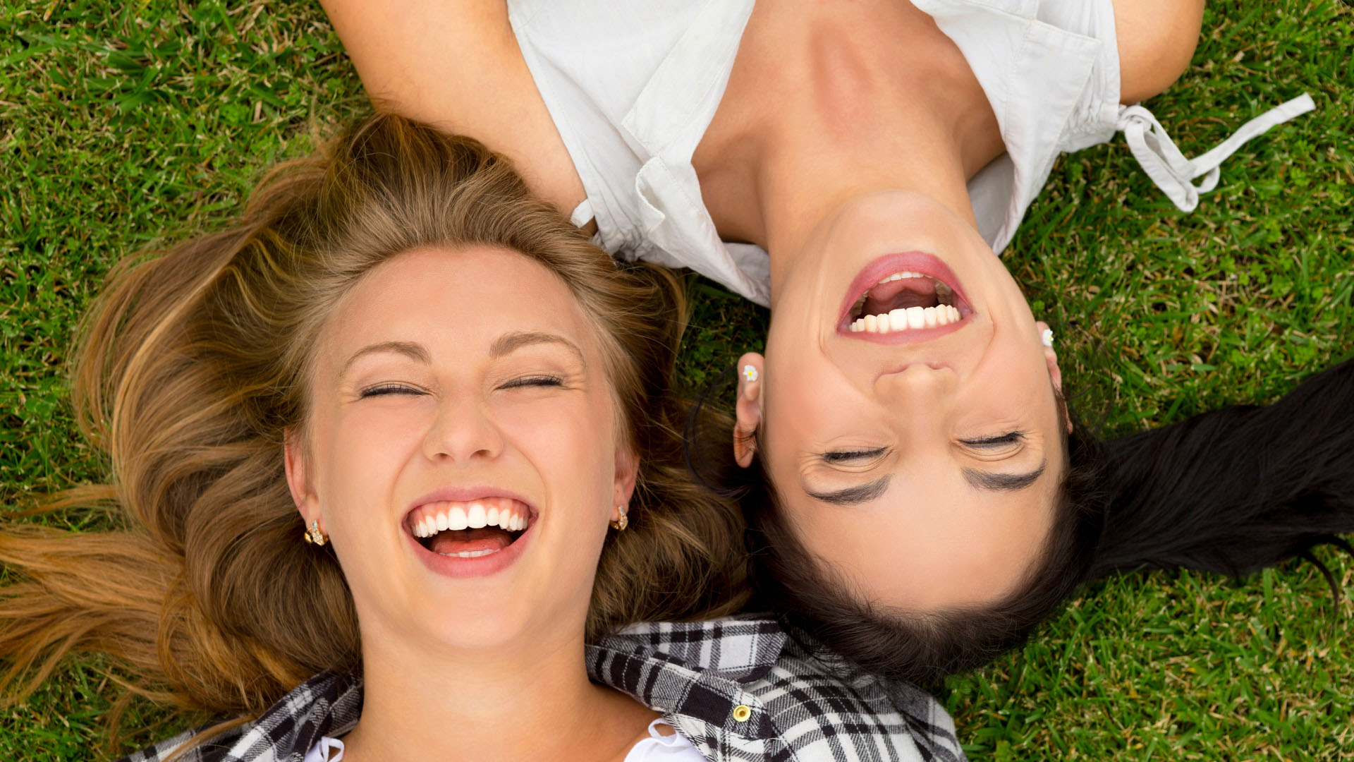 Two young women lying on grass laughing with bright white teeth, viewed from above