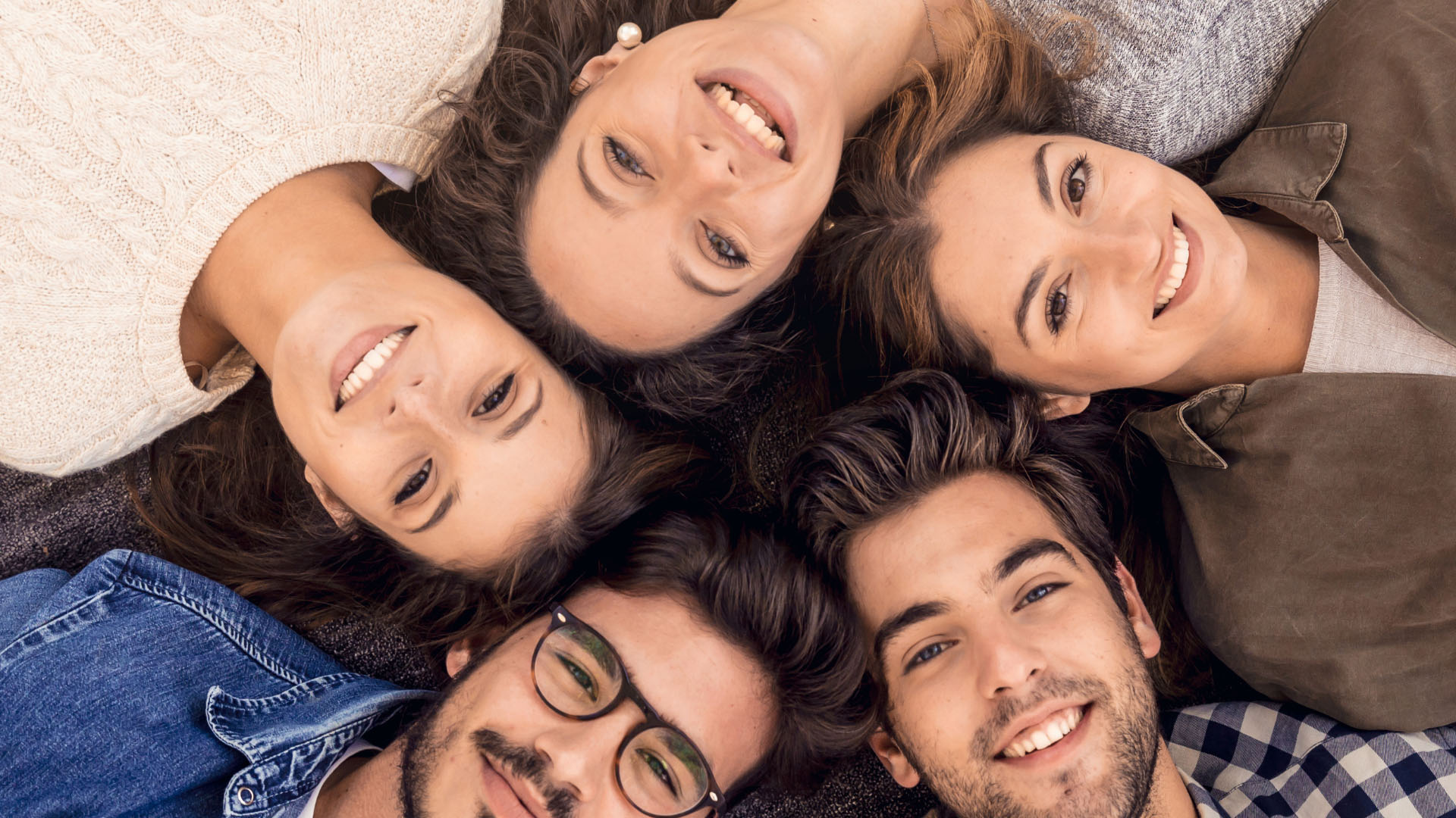Five smiling young friends lying on the ground with their heads together, looking up at the camera