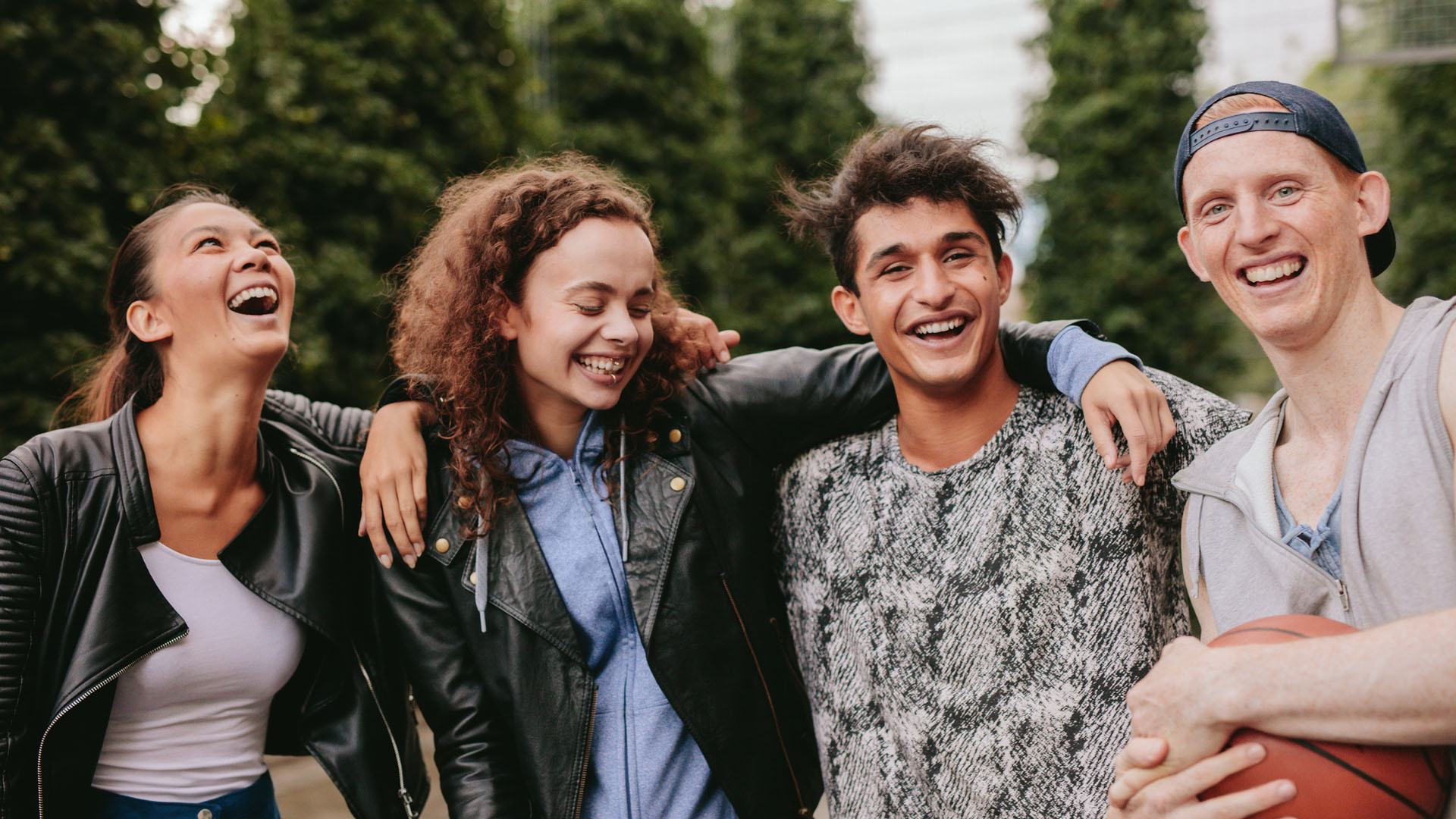 Four laughing teenagers with arms around each other outdoors, one holding a basketball, with trees in the background