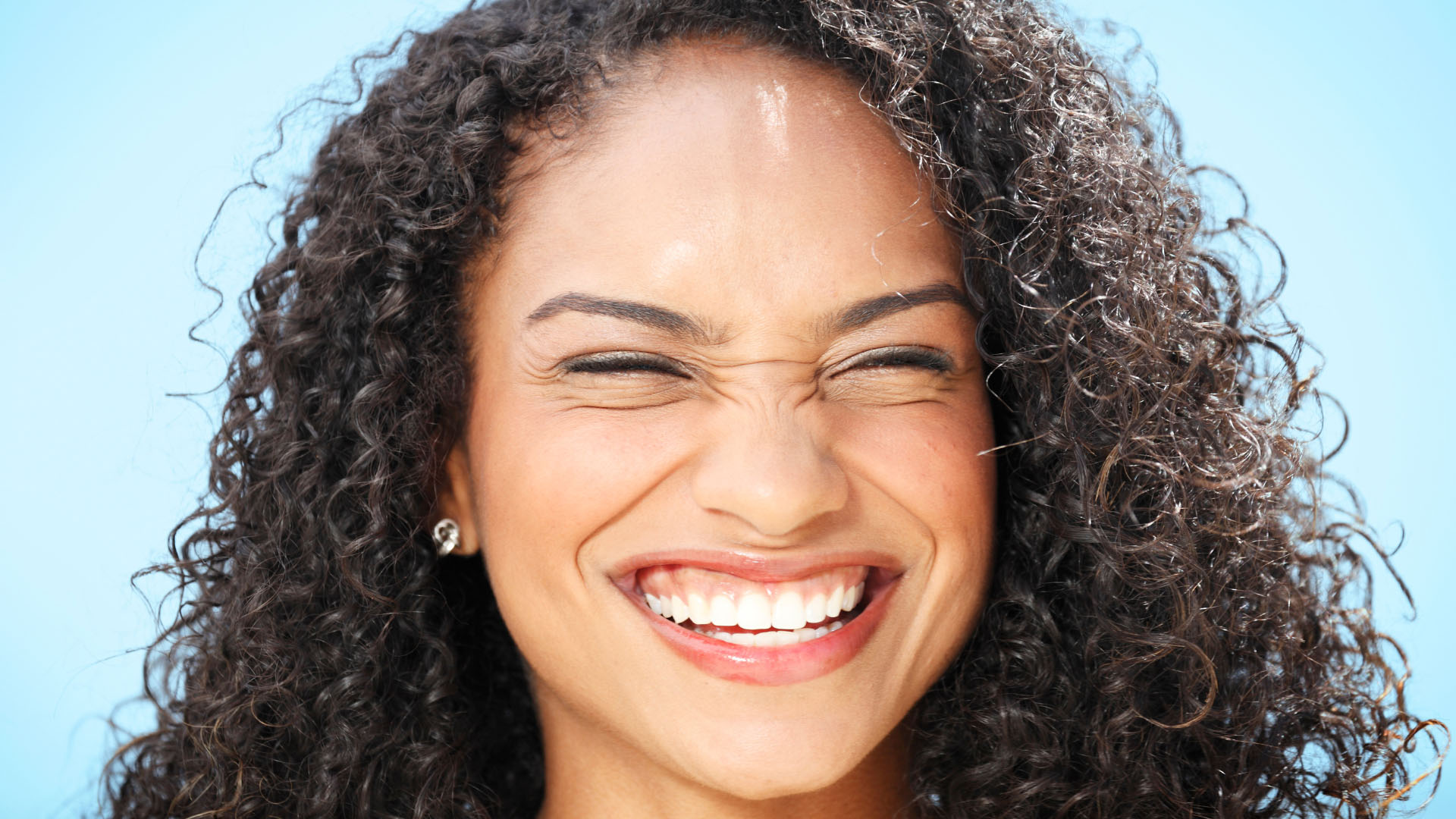 Woman with curly hair laughing broadly, showing white teeth, against a light blue background