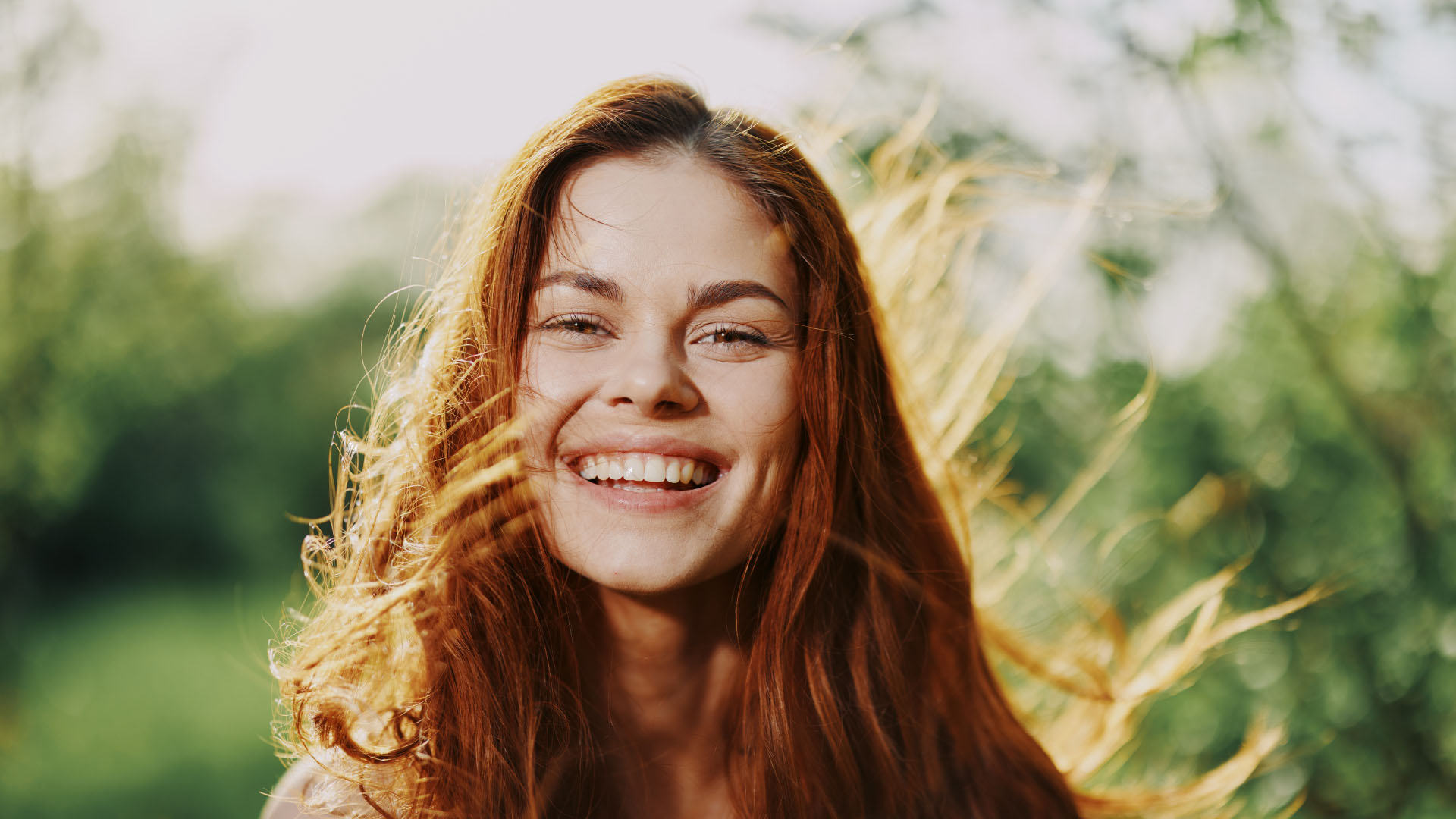 Young woman with long auburn hair blowing in the wind, smiling broadly outdoors against a blurred green background