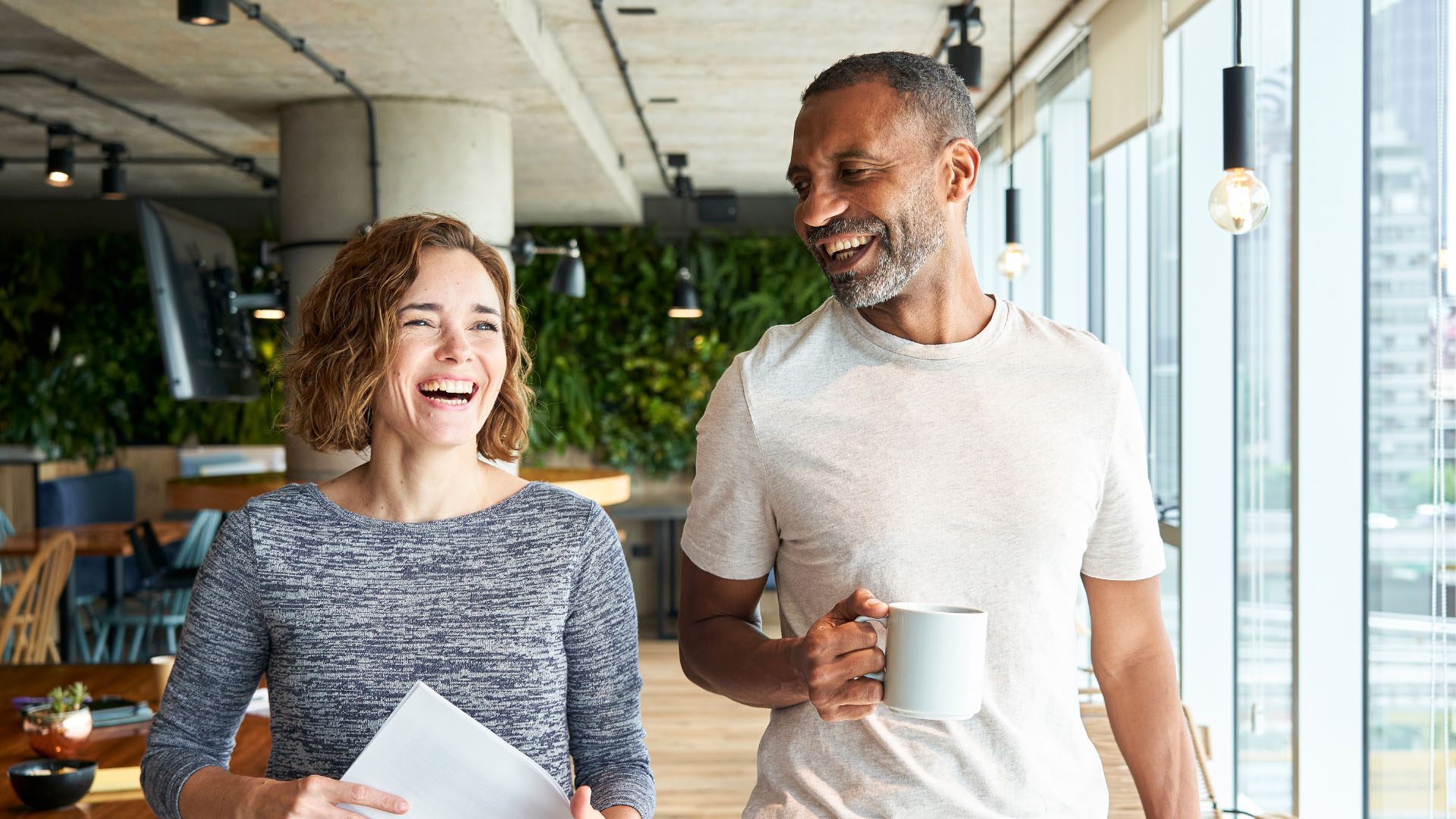 Two colleagues laughing together in a modern office, one holding documents and the other a mug, with a green plant wall behind them.