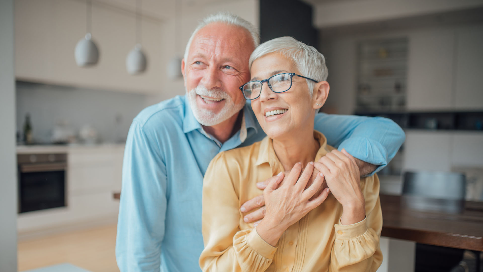 Smiling elderly couple embracing in a modern kitchen, the woman wearing glasses and a yellow blouse, the man in a light blue shirt
