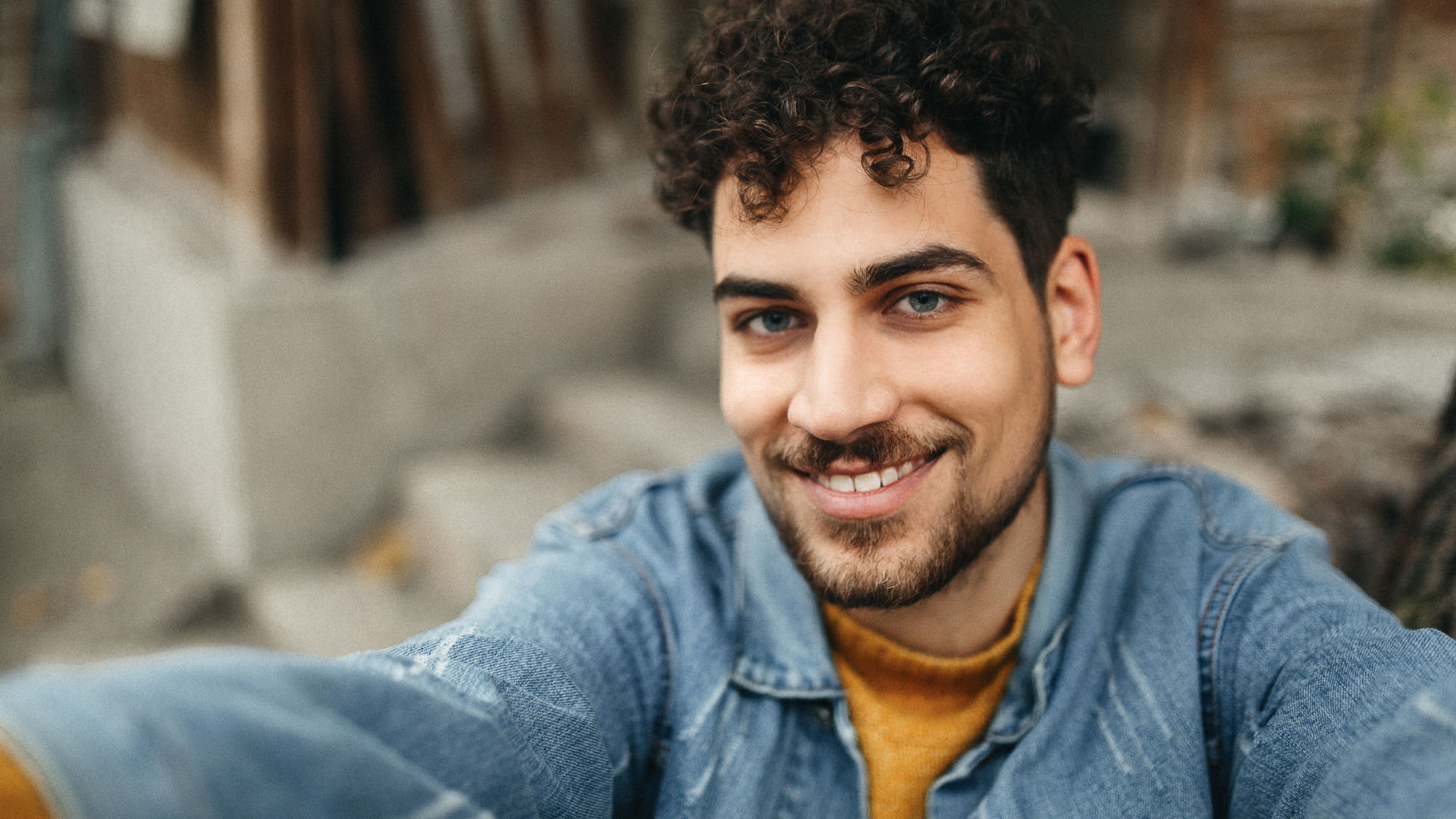 Young man with curly dark hair and a beard smiling outdoors, wearing a denim jacket over a mustard jumper.