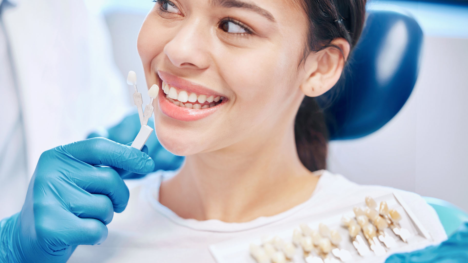 Smiling female patient in a dental chair as a gloved dentist holds a tooth shade guide against her teeth
