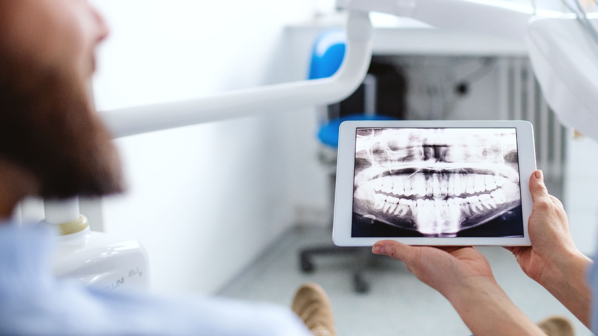 Dentist holding a tablet displaying a panoramic dental X-ray in front of a seated patient in a dental surgery