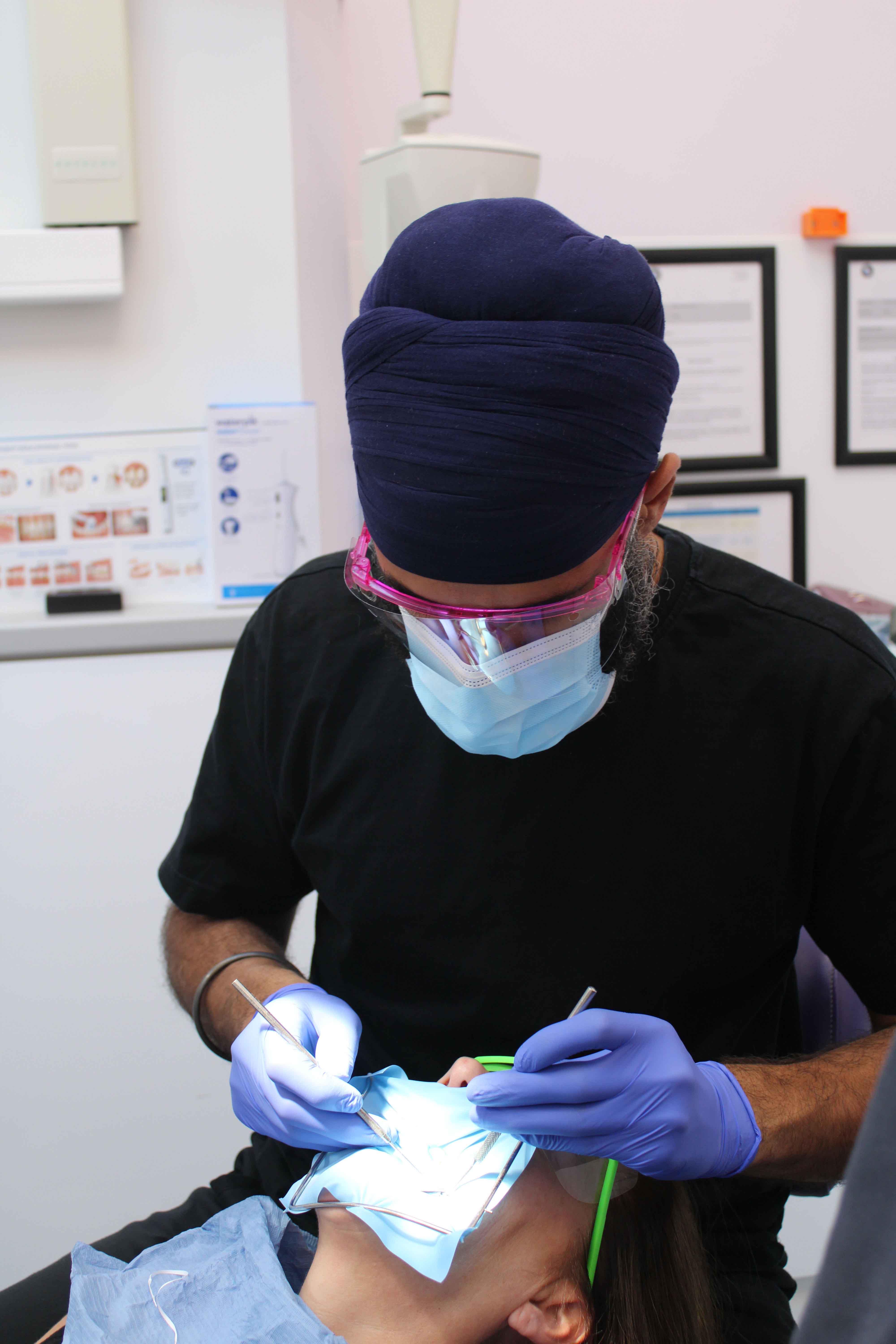 Dentist wearing a blue turban, mask and gloves examining a patient's mouth using dental tools in a clinic