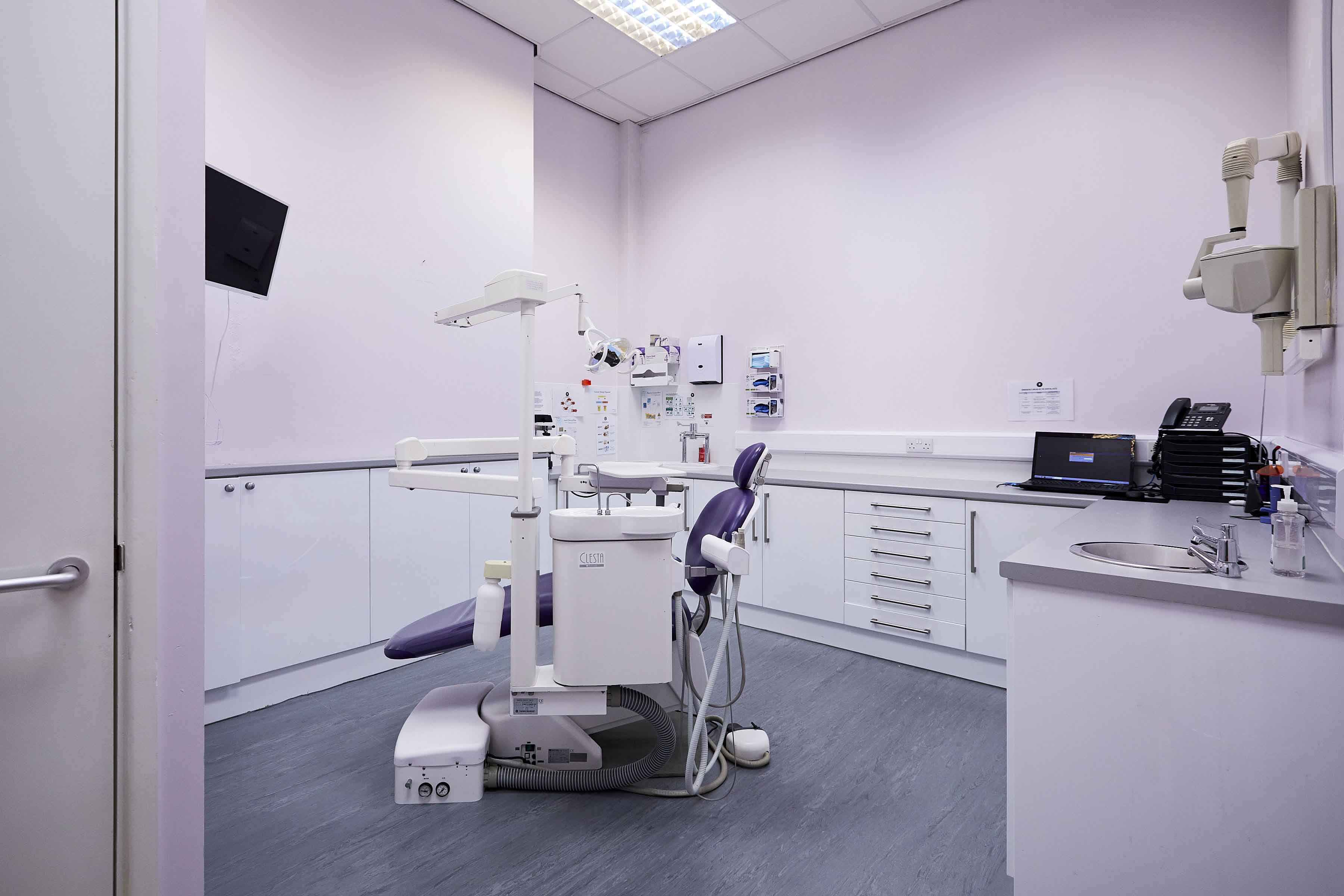 Clean dental treatment room with a purple patient chair, overhead light, X-ray equipment, and white cabinetry