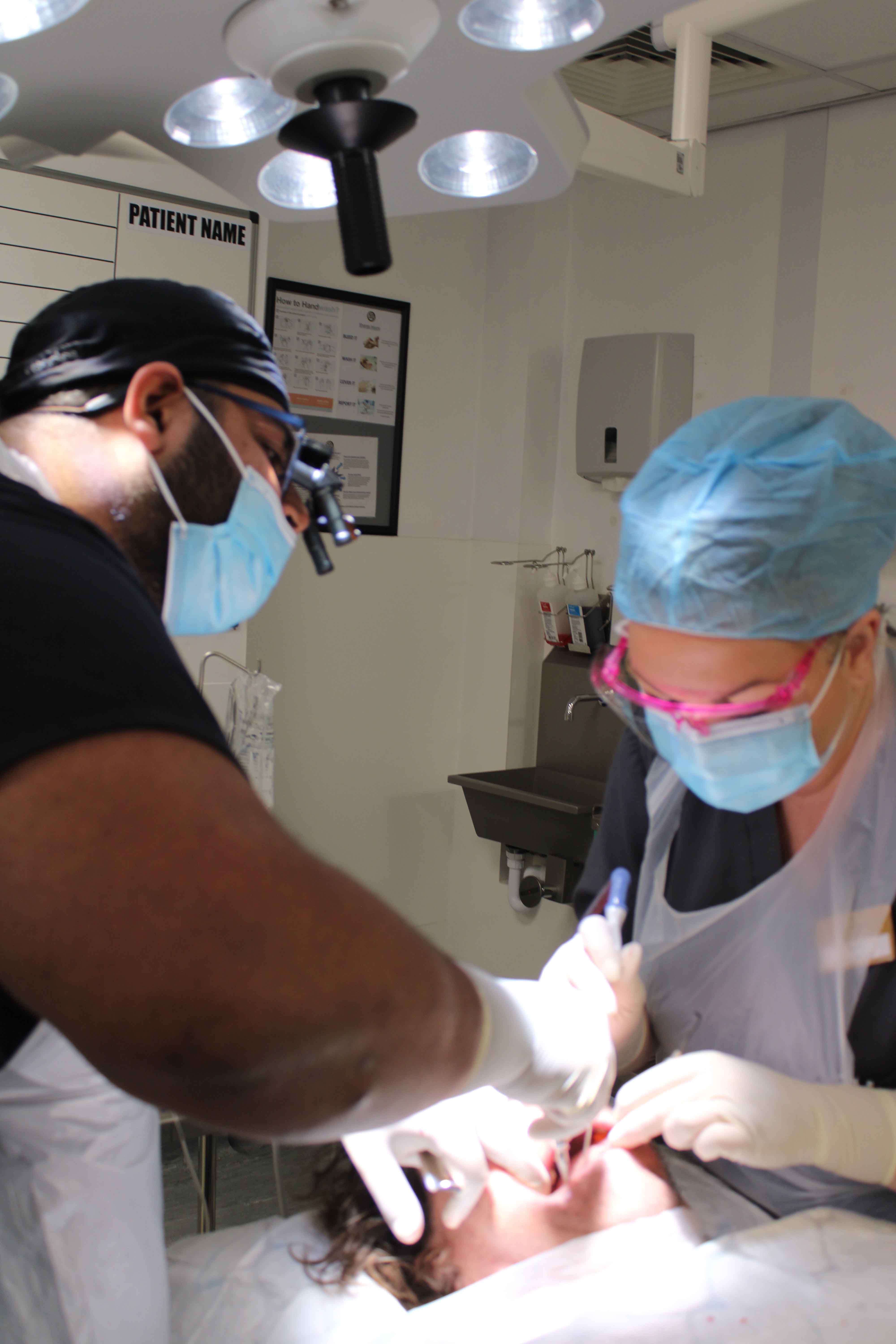 Two dental surgeons in masks and surgical caps performing a procedure on a patient under theatre lights