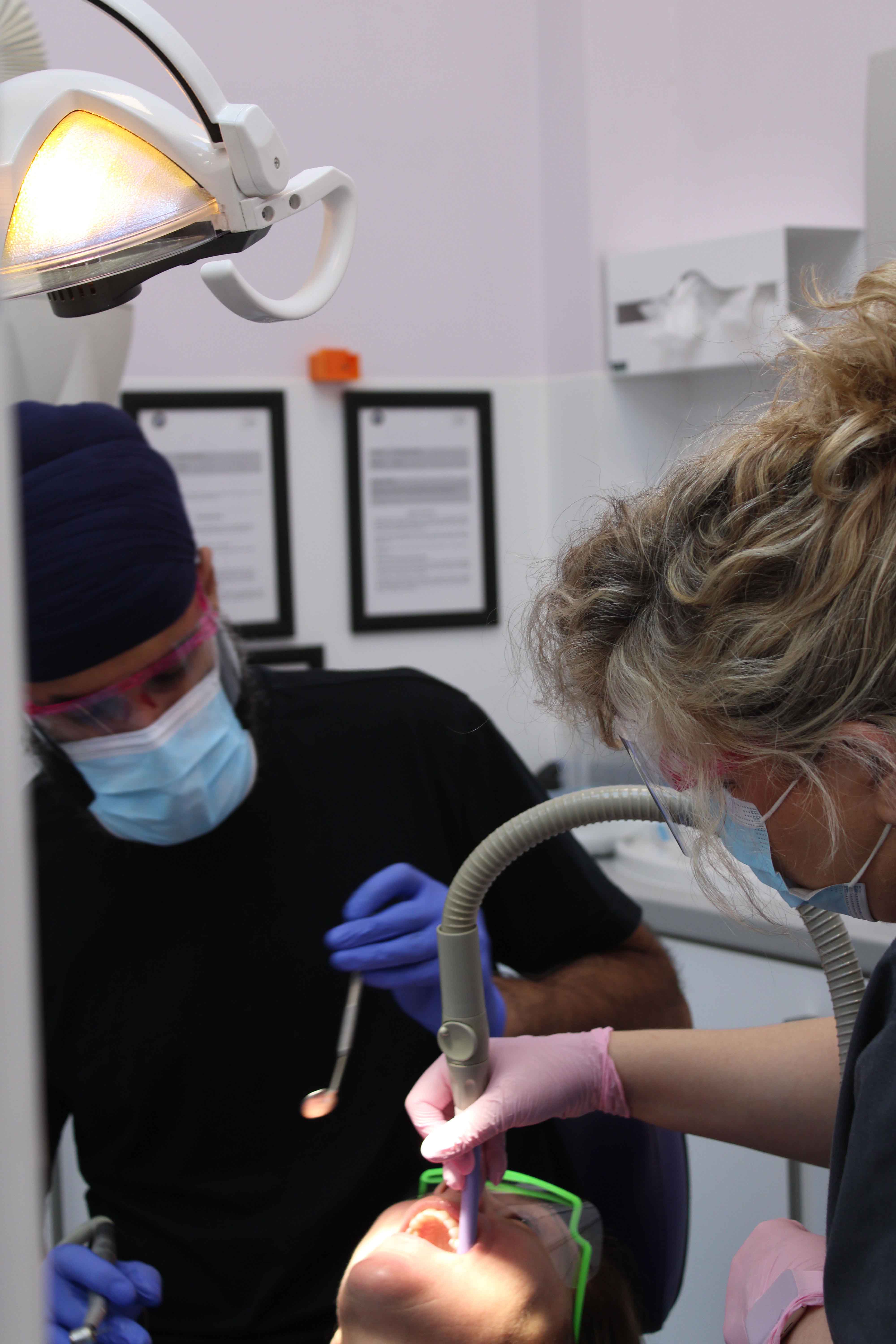 Two masked dentists wearing gloves treat a patient under a bright dental lamp in a clinical surgery