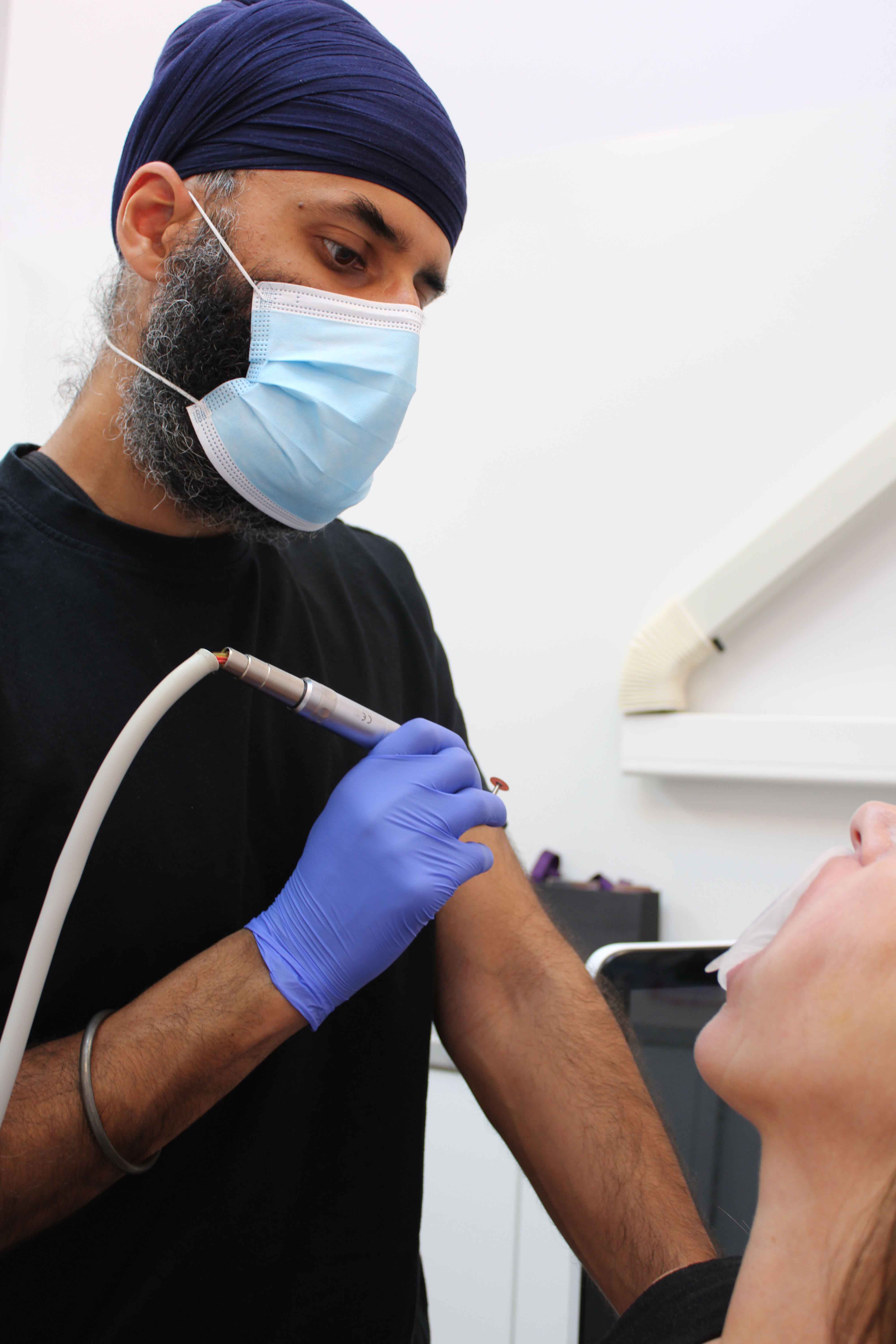 Bearded dentist wearing a blue turban and surgical mask treating a patient's teeth using a dental drill in a clinic