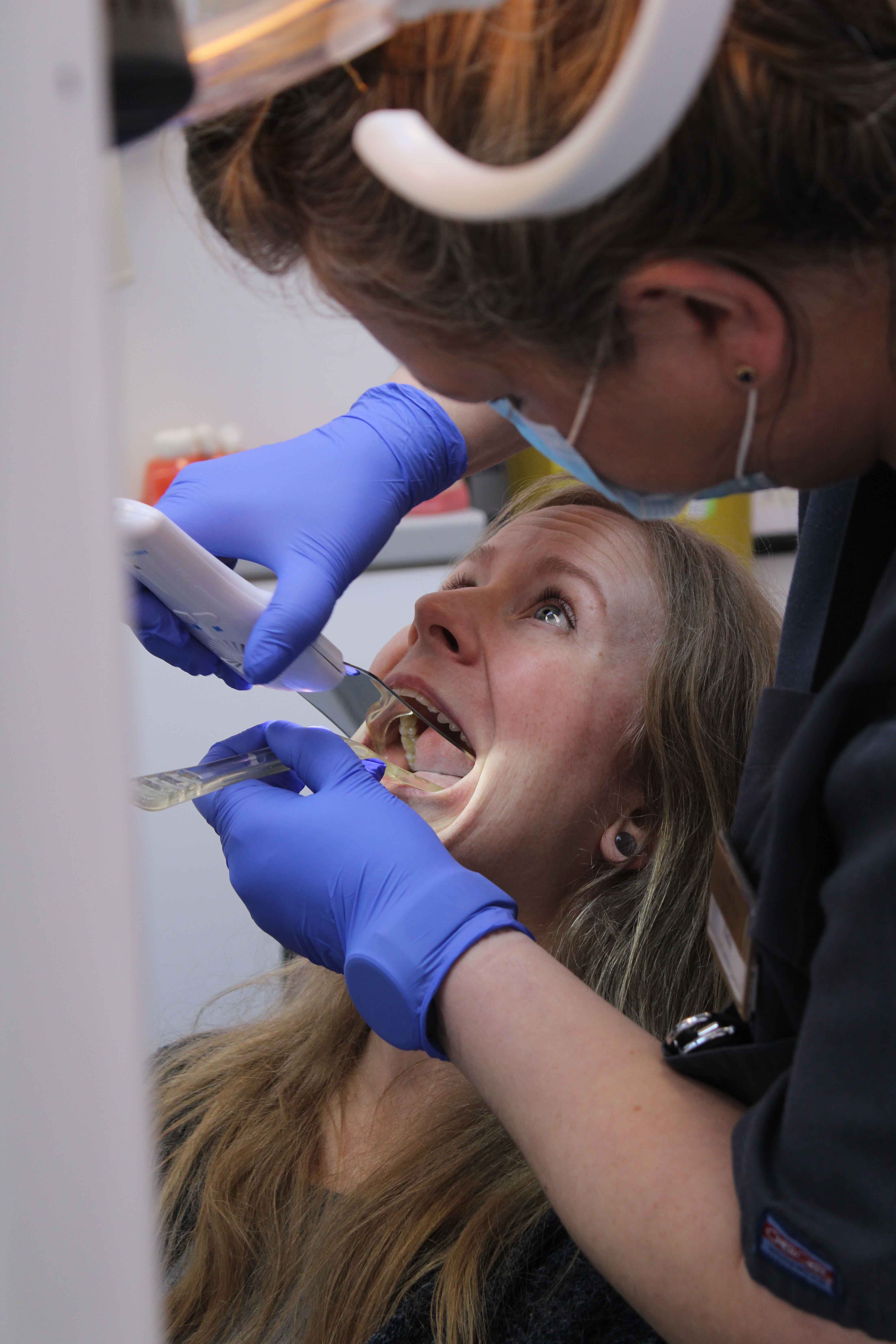 Dentist wearing blue gloves and a face shield examining a female patient's open mouth with dental instruments