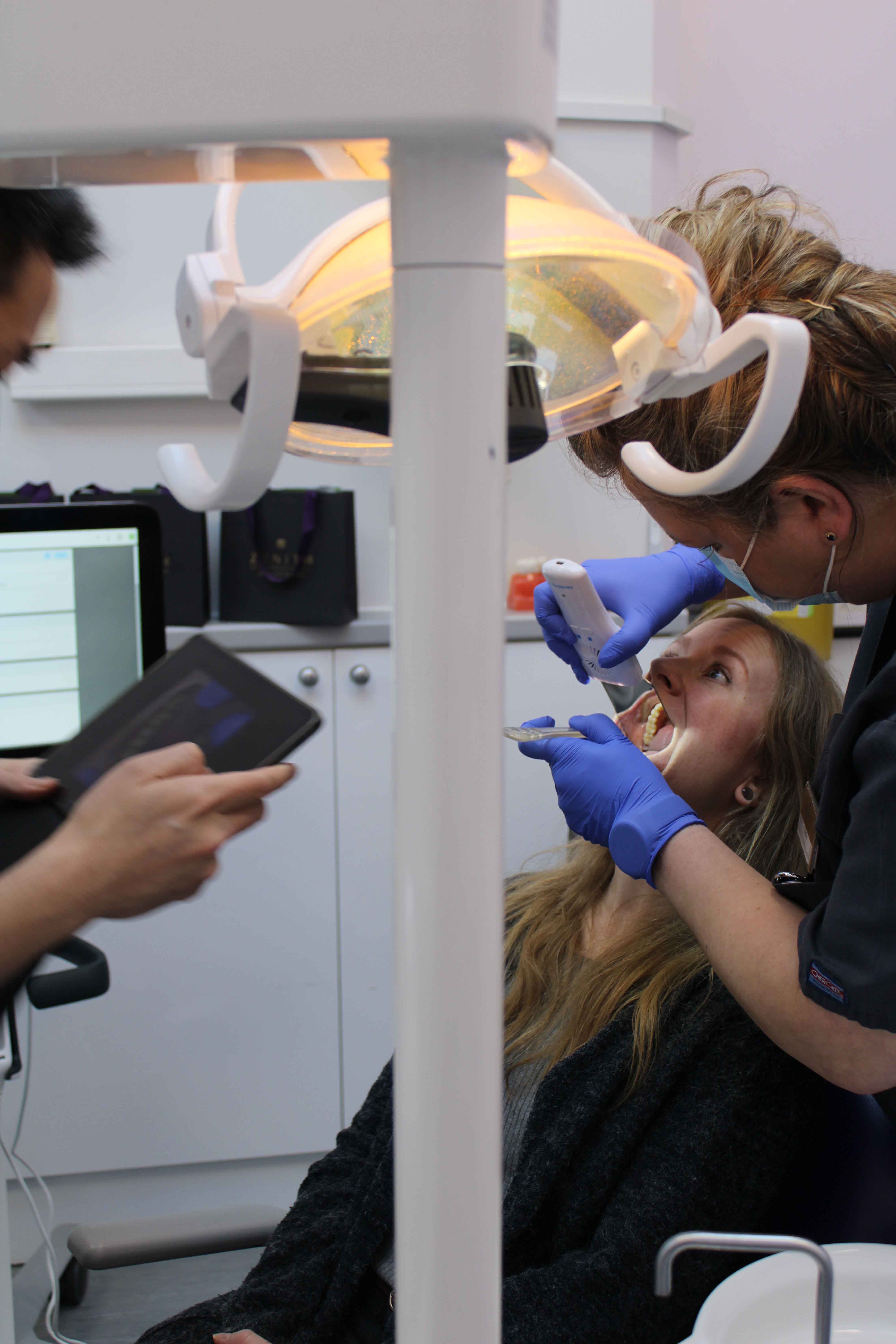 A dentist in blue gloves uses an intraoral scanner on a female patient reclined in a dental chair under a bright overhead light.