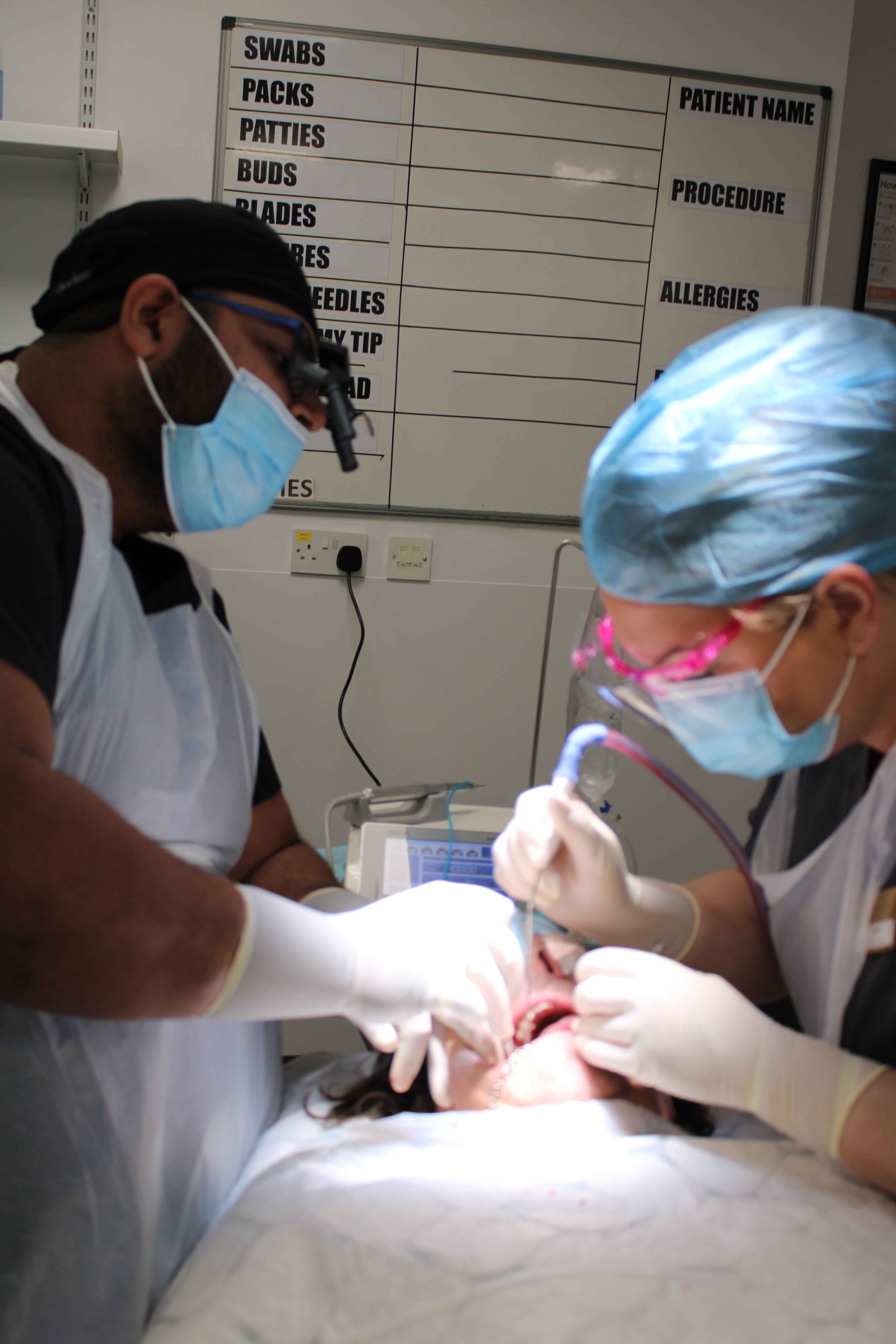 Two masked dental surgeons in gloves and scrubs performing a tooth extraction procedure on a patient in a clinical setting.