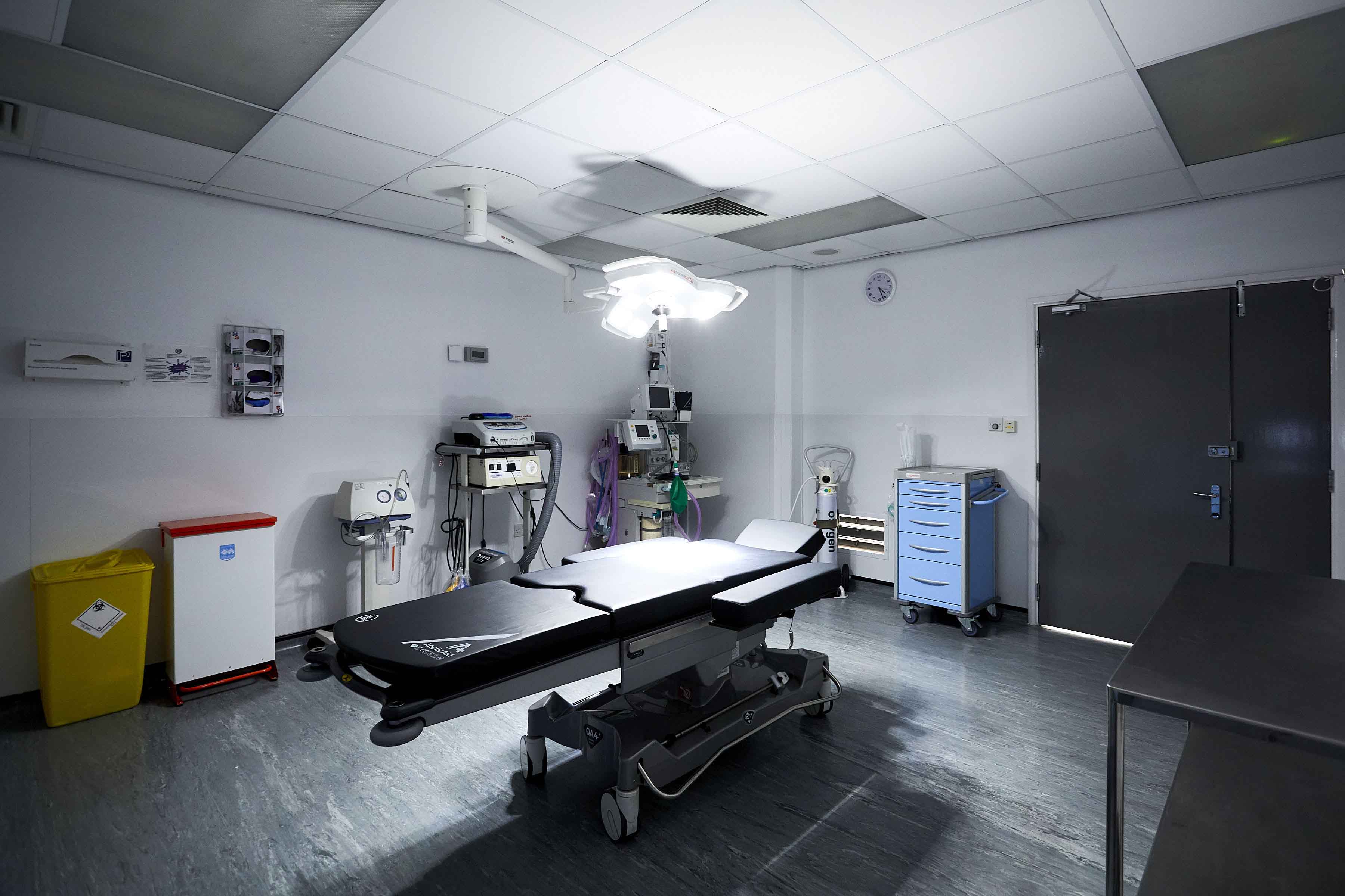 Empty hospital treatment room with a surgical table, overhead operating light, anaesthesia equipment, and medical trolley