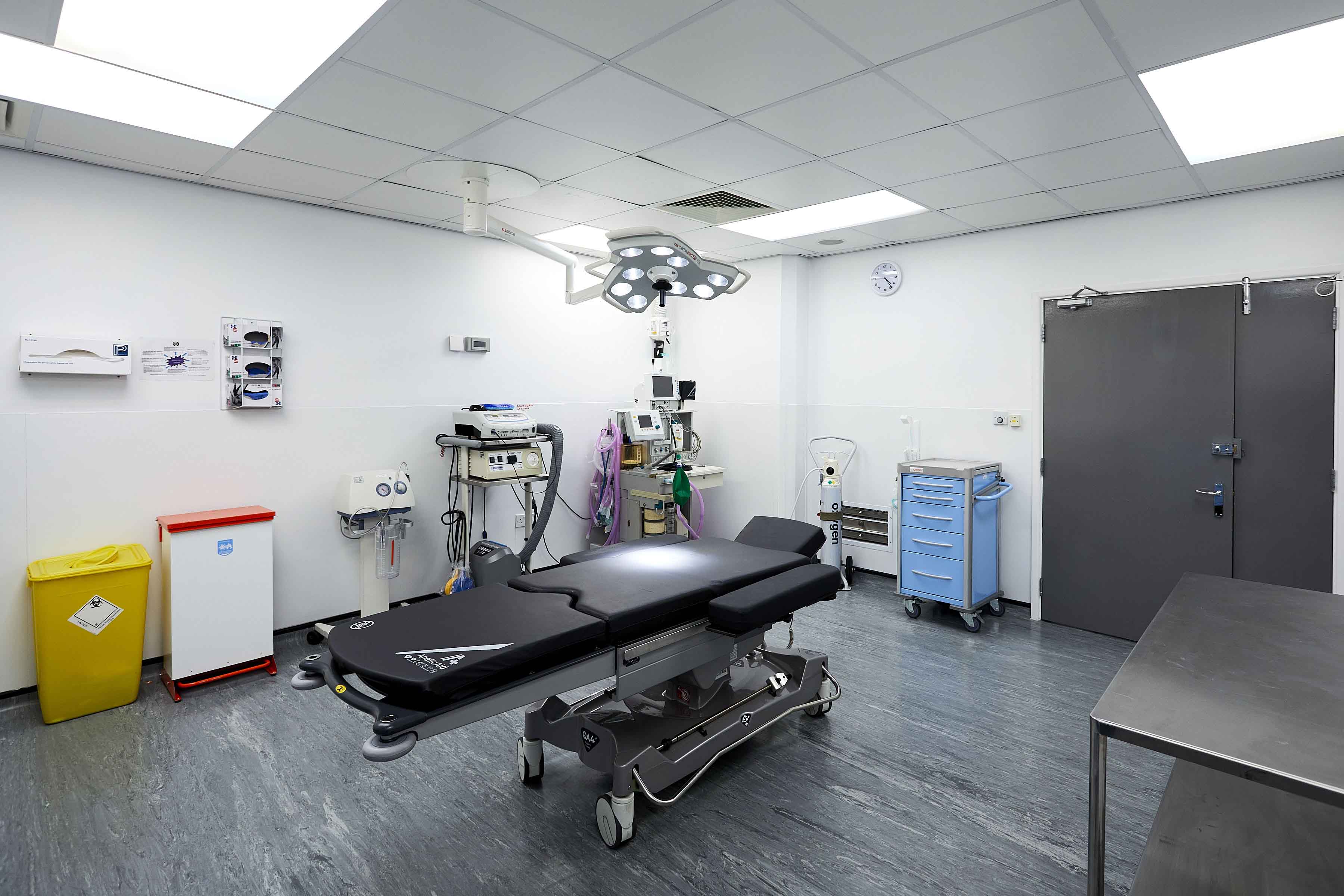 A clean surgical treatment room with a black operating table, overhead surgical light, medical equipment trolleys, and an oxygen cylinder.