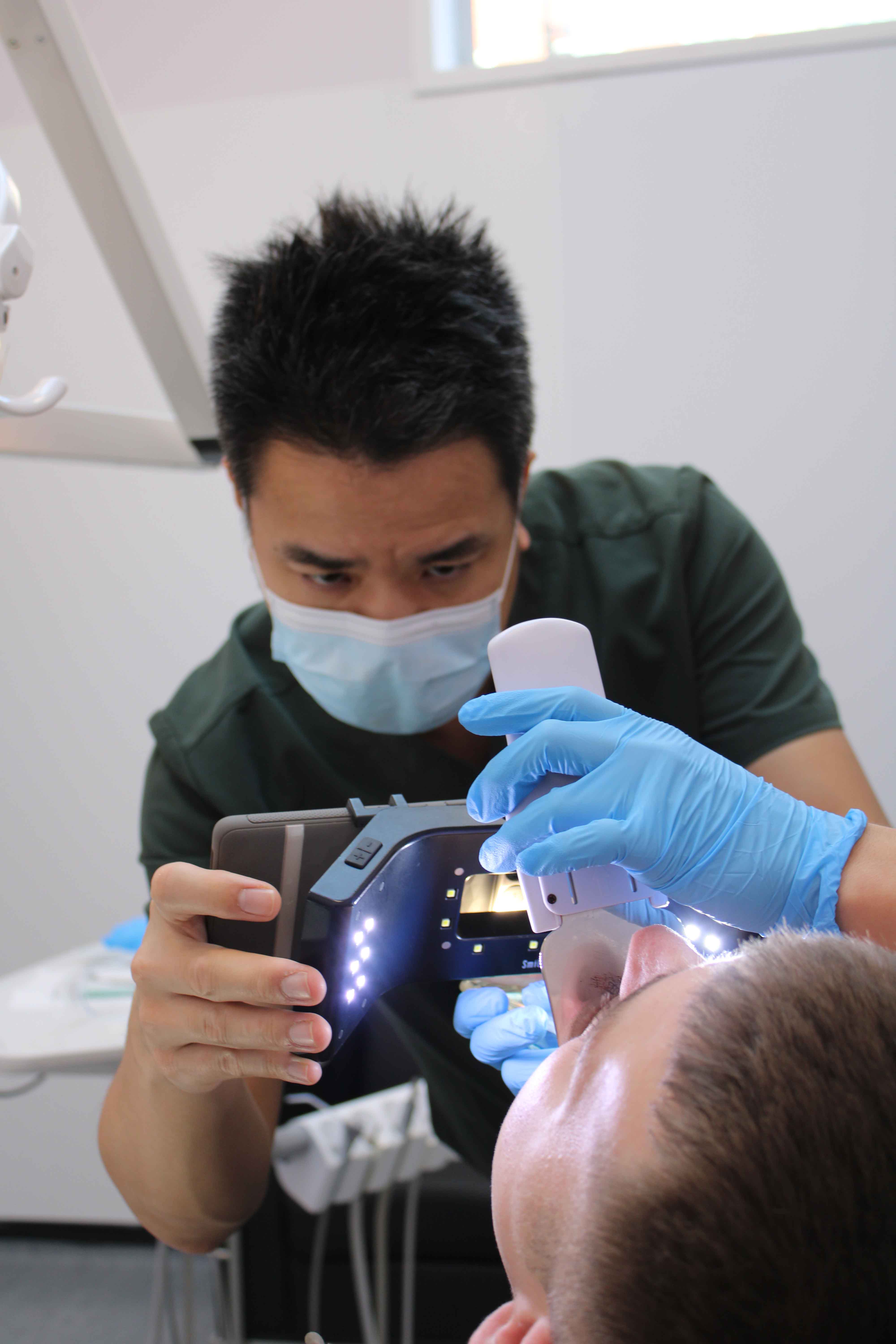 A dental professional wearing a face mask and blue gloves uses a handheld scanning device and illuminated equipment while examining a patient’s mouth.