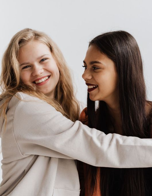 Two teenage girls laughing and hugging, one in a cream hoodie and one in a rust orange hoodie, against a white wall.
