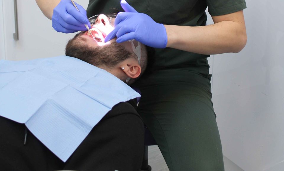 A dental professional wearing blue gloves examines a reclining patient’s mouth under a bright dental light.