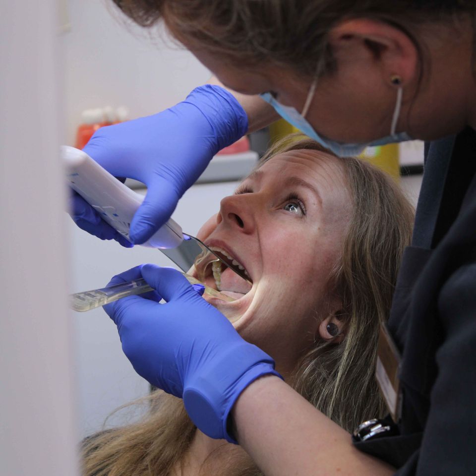 Dentist wearing blue gloves and a face shield examining a female patient's open mouth with dental instruments