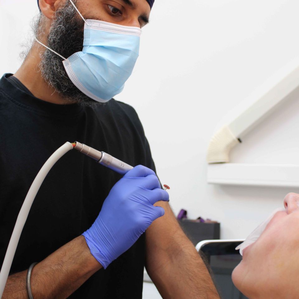 Bearded dentist wearing a blue turban and surgical mask treating a patient's teeth using a dental drill in a clinic