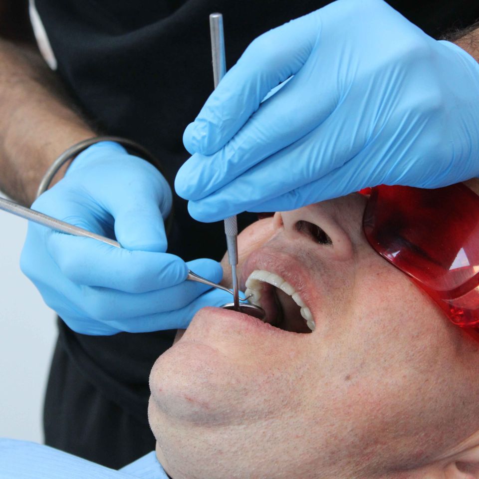 Dentist in blue gloves using dental tools to examine a male patient wearing red protective glasses