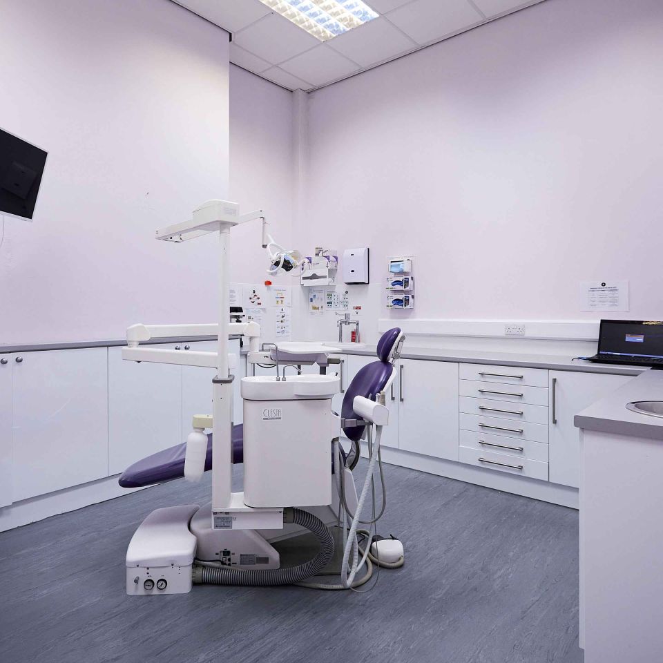Clean dental treatment room with a purple patient chair, overhead light, X-ray equipment, and white cabinetry