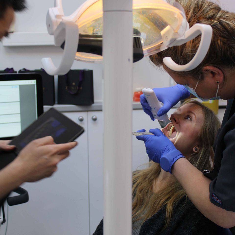 A dentist in blue gloves uses an intraoral scanner on a female patient reclined in a dental chair under a bright overhead light.