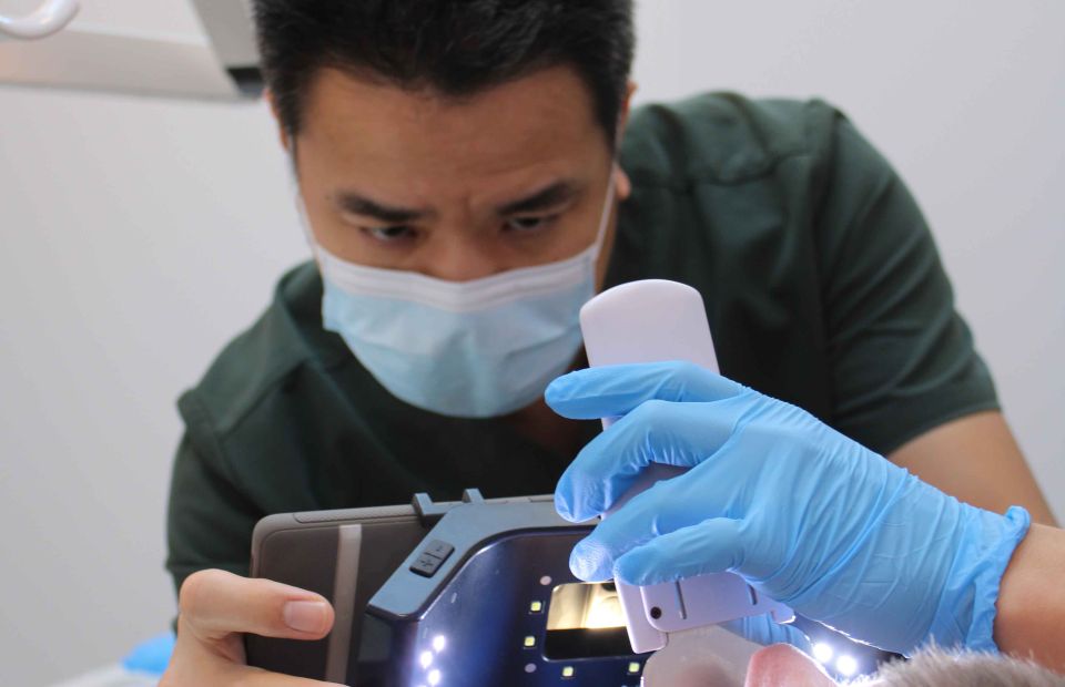 A dental professional wearing a face mask and blue gloves uses a handheld scanning device and illuminated equipment while examining a patient’s mouth.