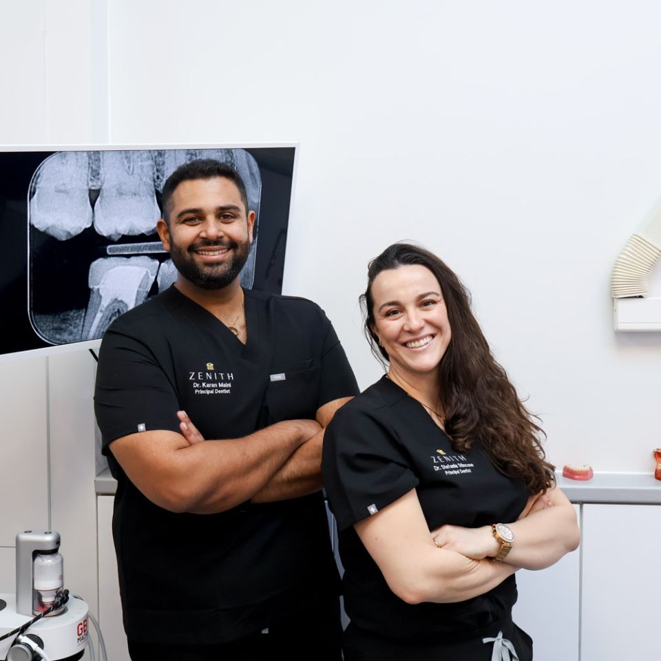 Two Zenith dentists in black scrubs smiling with arms crossed in a dental surgery, a dental X-ray displayed on screen behind them
