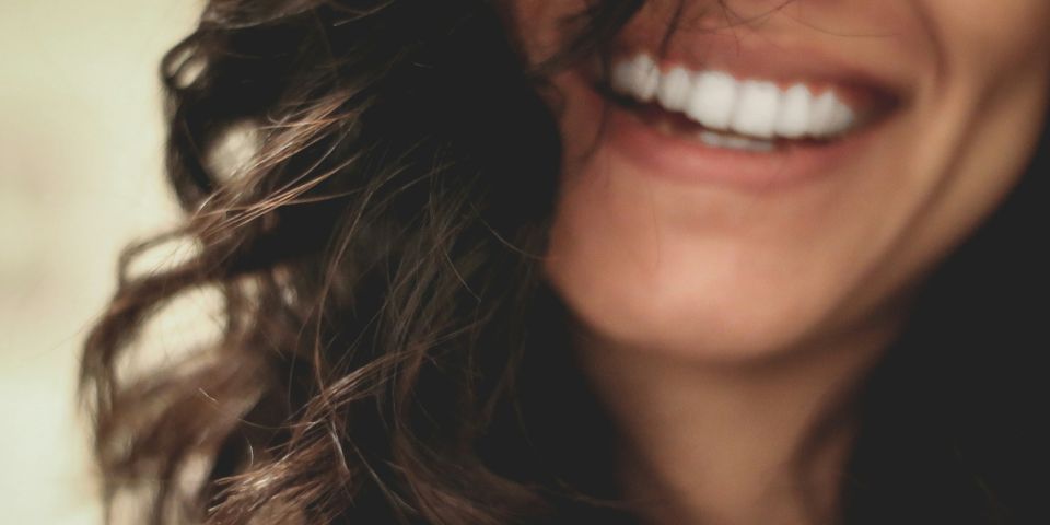 Close-up of a woman with dark curly hair laughing, showing bright white teeth against a soft blurred background