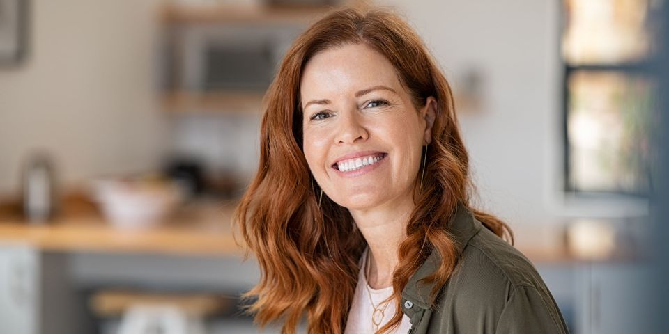 A middle-aged woman with long red hair smiling broadly, wearing a green shirt and gold jewellery, in a blurred kitchen setting.