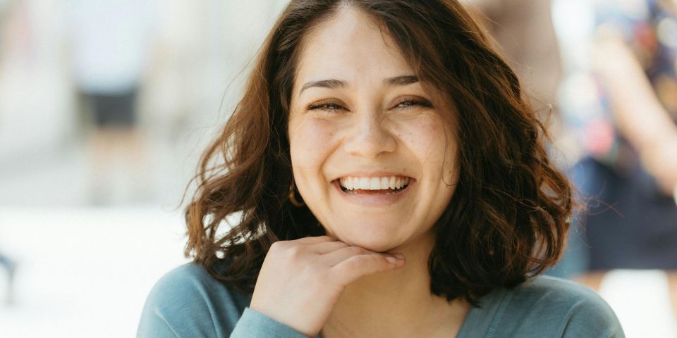 A young woman with curly brown hair laughing broadly, resting her chin on her hand, wearing a blue top outdoors.