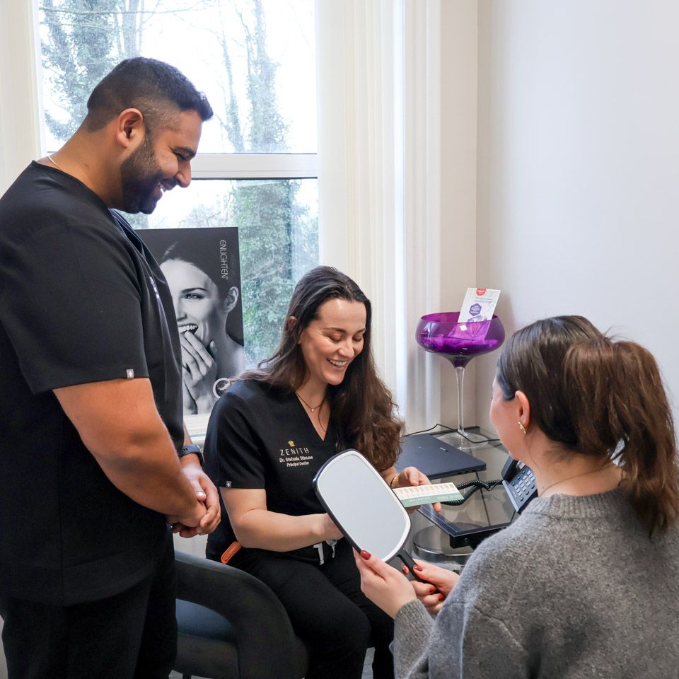 Two dentists in black scrubs smiling with a patient holding a hand mirror during a teeth whitening consultation at Zenith Dental