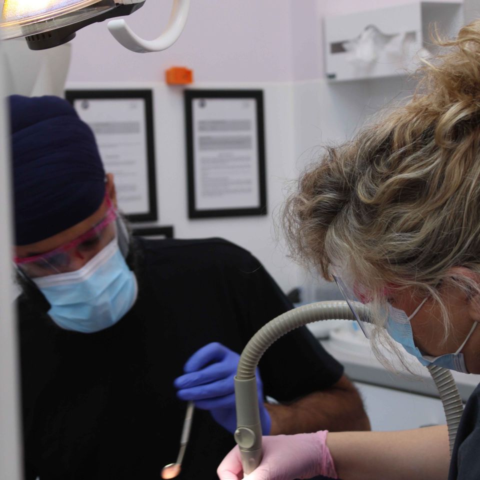 Two masked dentists wearing gloves treat a patient under a bright dental lamp in a clinical surgery